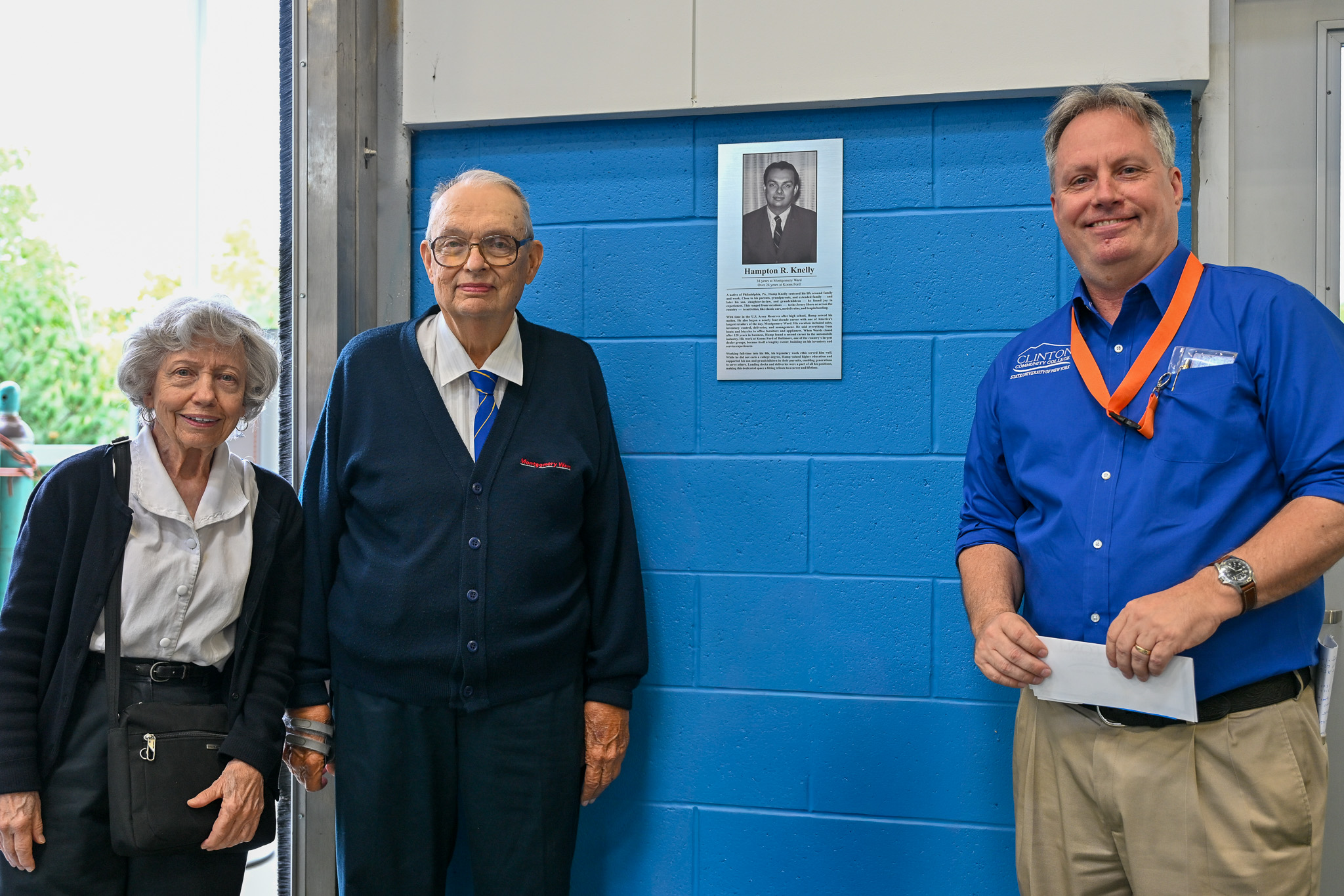 Three people standing next to a plaque with a picture and writing on it adhered to a blue cinder block wall