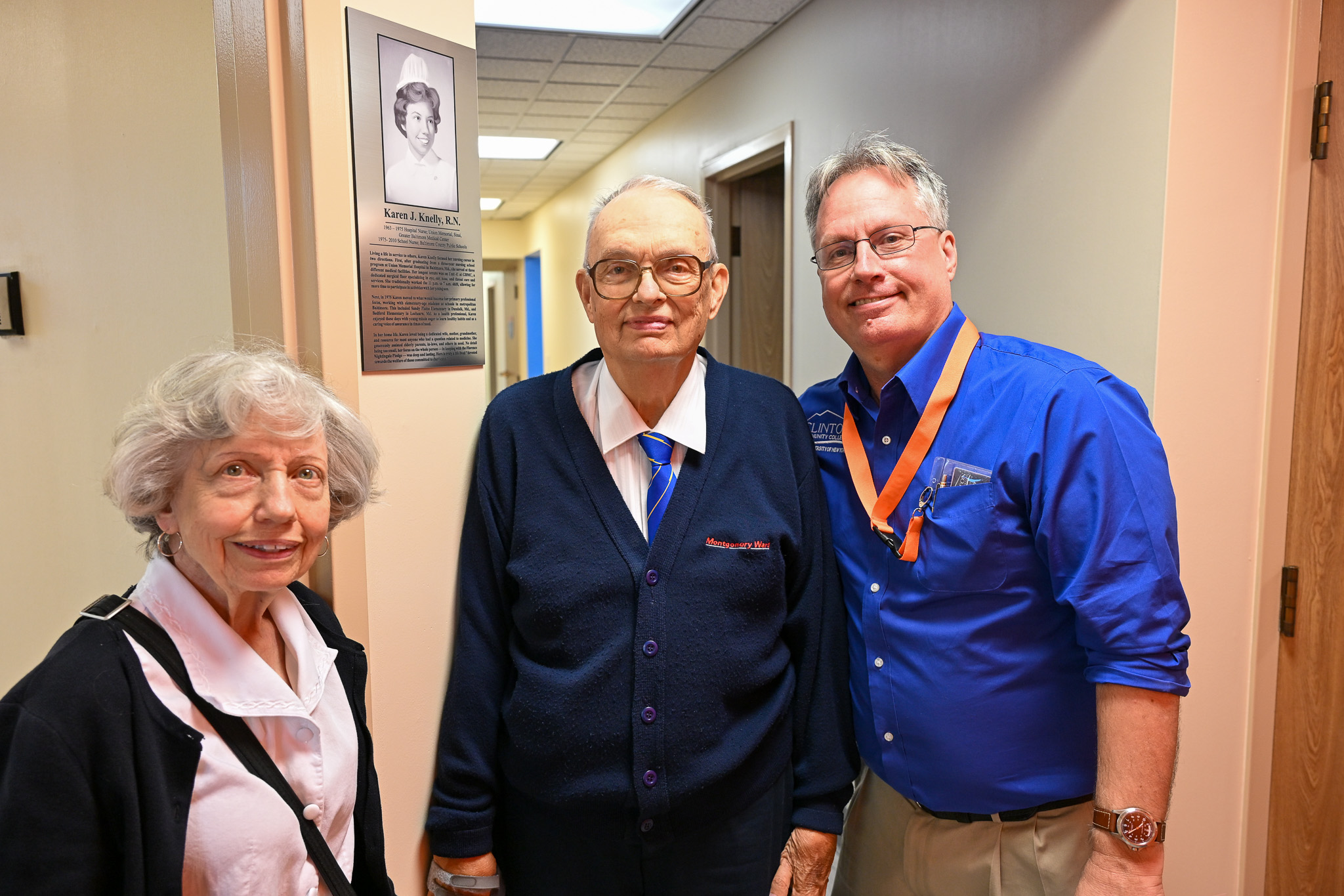 Three people standing next to a plaque with writing on it and a photo in an interior hallway