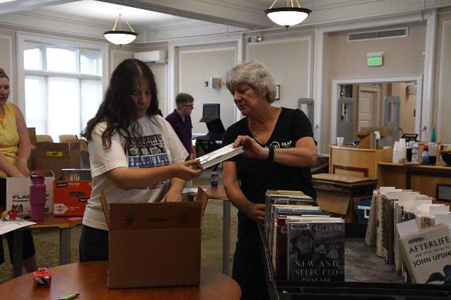 A person handing a hard cover book to another person as they pack a box