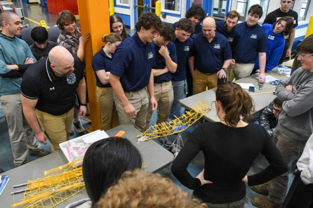 A group of students and teachers looking over two tables with a bridge made out of spaghetti spanning the opening between the tables
